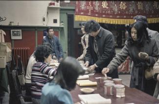 Photo couleur montrant des personnes d’origine chinoise dans une grande salle, certains sont assis derrière une table, à gauche, et d’autres sont debout de l’autre côté de la table, à droite.