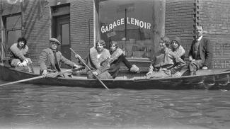 Cinq femmes et deux hommes sont dans une chaloupe dans une rue inondée devant un édifice de brique. 