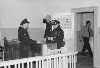 Photographie en noir et blanc montrant deux policiers en uniforme de chaque côté d’un comptoir. Un troisième policier est visible dans le corridor à travers le cadre d’une porte ouverte. 