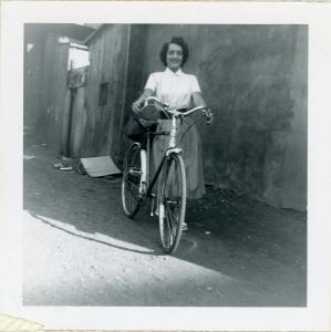 Photo en noir et blanc d’une jeune femme posant avec sa bicyclette dans une ruelle en terre, des hangars derrière elle.