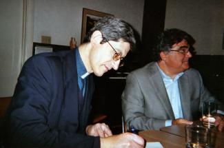 Photo couleur montrant deux hommes assis derrière une table. Celui à gauche signe un document.  