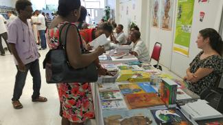 Dans une grande salle, des personnes se promènent d’une à une autre. Des livres sont disposés sur les tables et des personnes sont assises derrière la table.