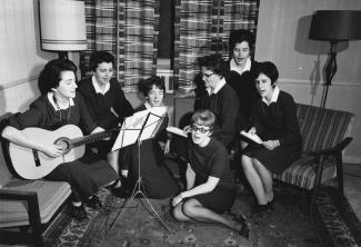 Sept femmes sont assises dans un salon sur des divans et sur le tapis. Une joue de la guitare et les autres regardent une partition pour chanter.