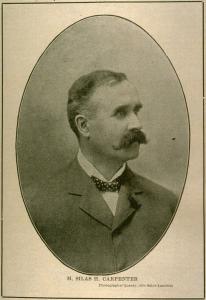 Photographie sépia, dans un médaillon, d’un homme blanc en complet et portant une moustache, sur un fond clair.