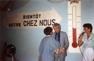 Une bannière à l’effigie d’un thermomètre est affichée au mur. Une femme, de dos, serre la main d’un homme. Une autre est debout et tient un microphone.