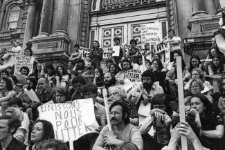 Une cinquantaine de personnes sont assisses sur les escaliers devant l’hôtel de ville de Montréal, pancartes à la main.