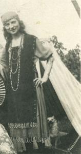 Jeune femme coiffée d’un foulard et portant une robe bohémienne et plusieurs colliers, devant une tente et un chien. 