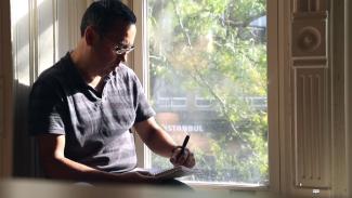 Un homme assis sur le bord d’une fenêtre écrit.
