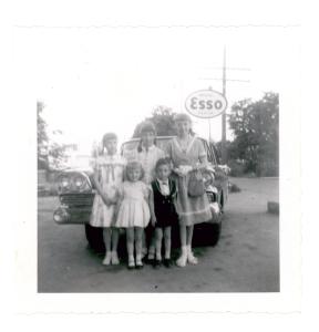 Photographie en noir et blanc montrant un groupe de cinq enfants d’âges variés bien habillés devant une voiture. Il y a un panneau de la compagnie ESSO à l’arrière à droite. 