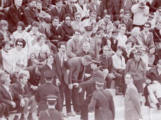 Photographie en noir et blanc montrant une foule dans des estrades. Deux jeunes personnes bien mises et portant des lunettes sont debout et crient. Des policiers les entourent et sont en mouvement vers le duo. 