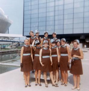 Onze hôtesses en uniforme (sans la veste) debout devant le pavillon du Québec.