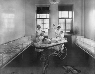 Photographie en noir et blanc montrant l’intérieur d’une pièce avec deux fenêtres au fond. Devant les fenêtres, deux infirmières tiennent des bébés dans leurs bras et un berceau se trouve devant elles. De chaque côté de la pièce, on peut voir des berceaux