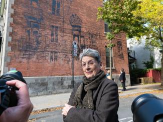 Une femme aux cheveux gris se tient devant un édifice dont un mur est en briques. Sur ce mur, le dessin d’une maison victorienne a été peint. 