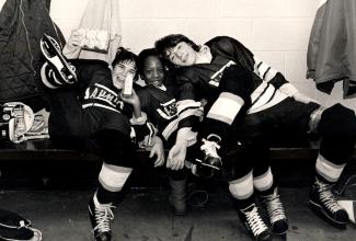 Trois jeunes hockeyeurs sont assis collés un sur l'autre sur un banc, dont un jeune Antillais au centre.