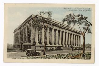 Carte postale en couleurs d’un grand immeuble à colonnade. Des voitures noires sont stationnées devant l’édifice et deux arbres feuillus sont visibles au premier plan. 