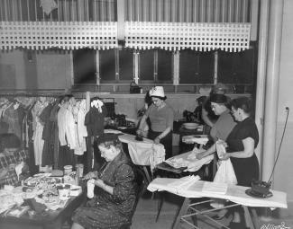 Dans une pièce où des vêtements sont suspendus, deux femmes reprisent des bas à une table; cinq femmes sont debout, quatre repassent, une s’est tournée vers sa voisine; une femme est assise dans le fond. 