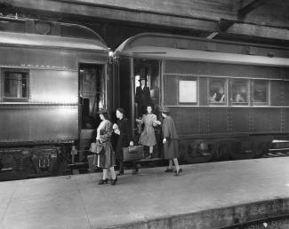 Deux wagons de train. Deux passagères marchent sur le quai avec leur valise. Une femme tend la main à une fille dans les escaliers du wagon, derrière elle, une fille est debout. Par une des fenêtres, on voit deux femmes sourire.