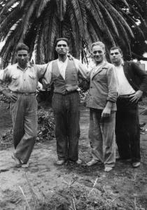 Photo en noir et blanc de quatre hommes, debout, devant un arbre. Un seul porte des chaussures. 