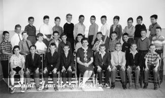 Photo en noir et blanc d’une classe de cinquième année. Il y a trois rangées d’enfants. L’enseignante est assis au milieu de la première rangée. 