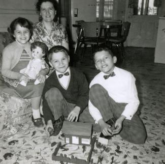 Uma mãe e os três filhos pousam com os presentes de Natal recebidos na sala. Vê-se a cozinha como plano de fundo.