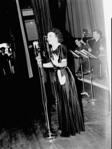 La chanteuse Alys Roby accompagnée par un orchestre sur la scène d'une salle de spectacle lors de la radiodiffusion de l'émission \"Tourbillon de la Gaieté\" de l'auteur Roger Marien par la station C.K.A.C. à Montréal.