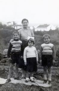 Photo en noir et blanc d’une mère et de ses trois enfants qui ont entre huit et cinq ans, dans une cour avec deux maisons en arrière-plan. 