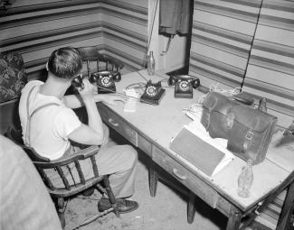 Un homme en tenue décontractée est assis derrière un bureau sur lequel il y a trois téléphones. L’homme utilise celui du centre.