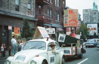 Deux voitures circulent sur une rue. Une remorque est attachée à l’une d’elles. Des affiches sont placardées sur une voiture et sur la remorque. Un homme tient une affiche dans la remorque. Un homme et des enfants sont sur le trottoir. 