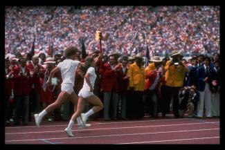 Photographie des deux relayeurs, vêtus de blanc, qui courent sur la piste en tenant la torche olympique.