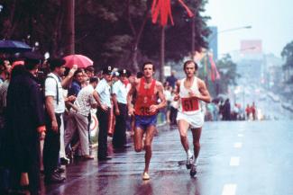 Photographie de deux athlètes courant dans la rue sous la pluie, avec une foule alignée sur le côté.