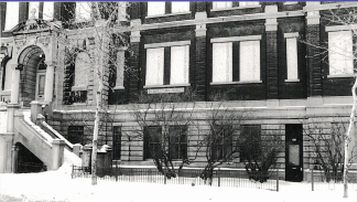 Photo noir et blanc montrant une partie de la façade d’un édifice ancien ayant abrité une école. On peut y lire l’inscription « Chamilly-de-Lorimier ». Les fenêtres des étages sont placardées.