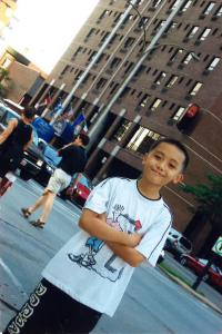 A young boy with his arms crossed, standing in a Montréal street. A high-rise hotel is visible in the background.