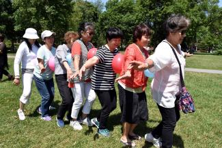 Seven women walk one behind the other in a park, balancing balloons between themselves without using their hands. 