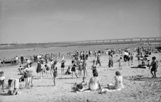 Plage avec plusieurs dizaines de baigneurs de tous âges, dont certains sont dans l’eau du fleuve, avec au loin le pont Victoria.