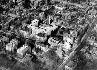 Photographie en noir et blanc montrant une vue aérienne d’un secteur densément habité. Un peu excentré, se trouve un large bâtiment en forme de H avec une longue cheminée cylindrique située à l’arrière de l’édifice.