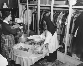 Deux femmes dans une petite pièce, vestiaire de vêtements, table avec légumes et pain et chaudron. 