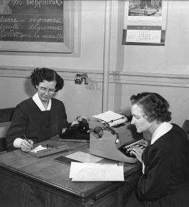 Deux femmes sont assises une face à l’autre à un bureau de travail. Une tape à la dactylo et l’autre prend des notes à la main et a une main posée sur le combiné du téléphone. 