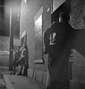 Deux femmes sont appuyées sur un mur de brique et sont éclairées par la lumière d’un lampadaire électrique. Un homme, de dos, les regarde. 