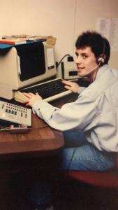 Photo couleur de 1990 montrant un homme assis à son bureau de travail, avec un casque d’écoute avec micro, et sur la table devant lui, un ordinateur et un téléphone. 