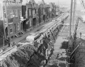 Photo en noir et blanc d’une rangé de maisons du côté gauche d’une rue et du côté droit, travaux de construction montrant une longue tranchée. 