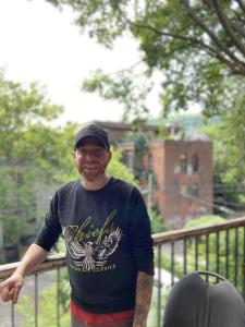 Un homme souriant habillé d’un chandail et d’une casquette noirs se tient debout sur un balcon en hauteur.