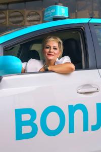 Photo couleur format portrait d’une chauffeuse de taxi souriant au photographe par la fenêtre ouverte de la portière. 