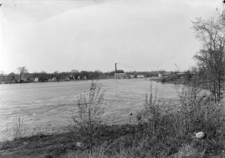 Rivière bordée par des berges couvertes de quelques arbres et arbustes avec au loin un barrage hydroélectrique et divers autres bâtiments.