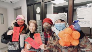 Photo couleur montrant quatre femmes d’origine chinoise dans une salle, l’une tient un sac d’oranges et deux tiennent des cartons rouges avec un texte en chinois.