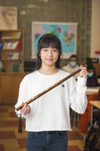 Photo d'une jeune fille d'origine chinoise tenant un objet de forme longue en bois dans une classe. L'arrière-plan est flou. 