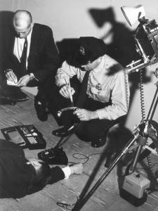 Photographie en noir et blanc, en plongée, montrant un policier passant un pinceau sur un combiné de téléphone.