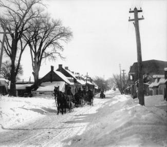 Photographie en noir et blanc représentant une série de petites maisons bordant un chemin de campagne, recouvert de neige.