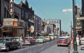 Photo couleur de la rue Sainte-Catherine à partir de la rue Saint-Dominique dans les années 1950 avec des voitures et les édifices commerciaux de chaque côté et leurs enseignes.