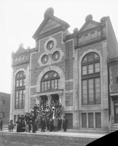 Photographie en noir et blanc d’une synagogue. Au centre de la façade se trouvent trois fenêtres circulaires à l’intérieur desquelles est visible une étoile de David. Un groupe de personnes se tient sur le parvis du bâtiment.