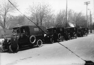 Photographie en noir et blanc où on voit des camions de type fourgon cellulaire avec le mot police inscrit sur le côté. Un policier se tient devant la rangée de véhicules. 